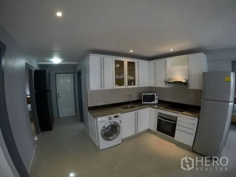 Condominium for rent in Watthana, Bangkok - Modern L-shaped kitchen with white cabinets, oven, cooktop, washing machine and fridge.