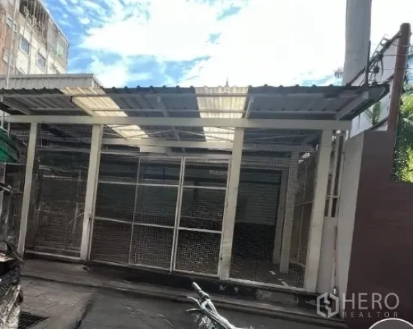 Retail Space for rent in Khlong Toei, Bangkok - Street-front single-storey retail unit with metal mesh facade, roller shutter doors, and a corrugated roof with skylight panels in Khlong Toei, Bangkok.