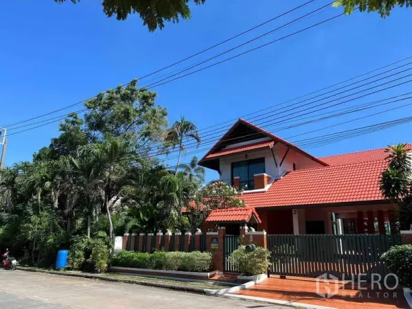 独立式住宅 出租在Bang Phli, Samut Prakan - 红瓦屋顶独栋别墅外观，木栅栏与茂密绿植，蓝天下。