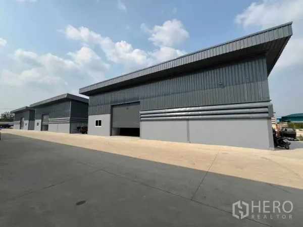 货栈 出租在Pathum Thani - 一排钢板外墙仓库沿宽阔通道排列，天空多云。