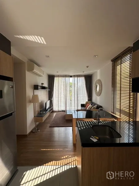 Condominium for rent in Watthana, Bangkok - Open-plan living room with wood floors, kitchen island, and floor-to-ceiling curtains leading to a sunny balcony.