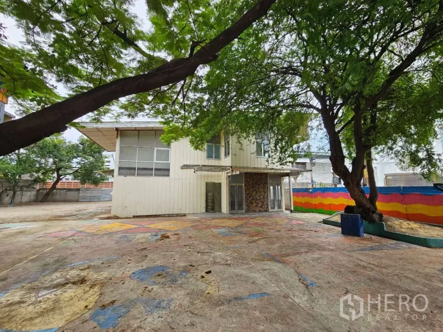 Detached House for rent in Watthana, Bangkok - Two-storey detached house with a large paved yard, mature shade trees, and a colorful boundary wall.