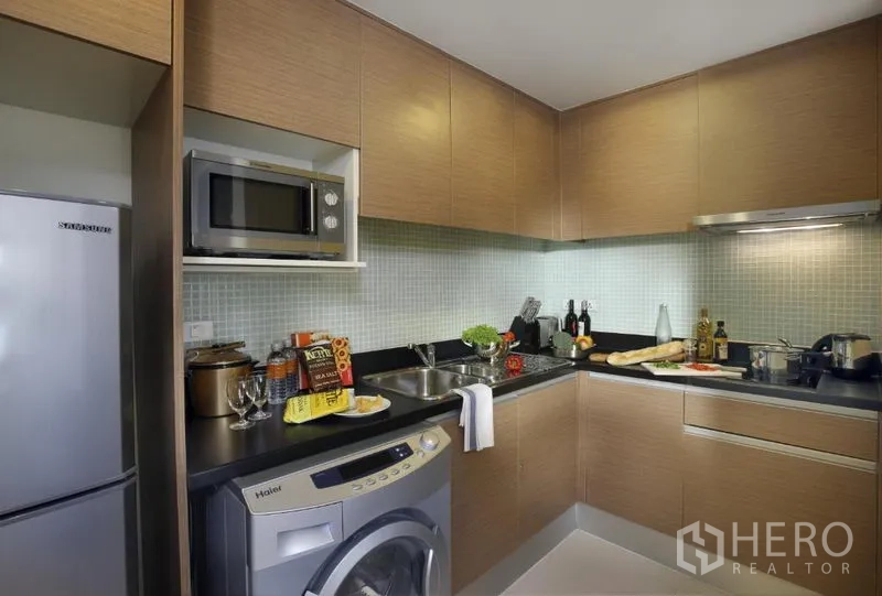Condominium for rent in Khlong Toei, Bangkok - Modern L-shaped kitchen with wood cabinets, microwave, fridge and in-unit washing machine at a Sukhumvit condo.