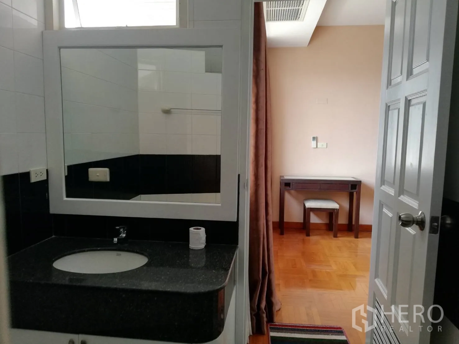 Condominium for rent in Watthana, Bangkok - Bathroom vanity with granite countertop and big mirror, looking out to a room with desk and curtains.