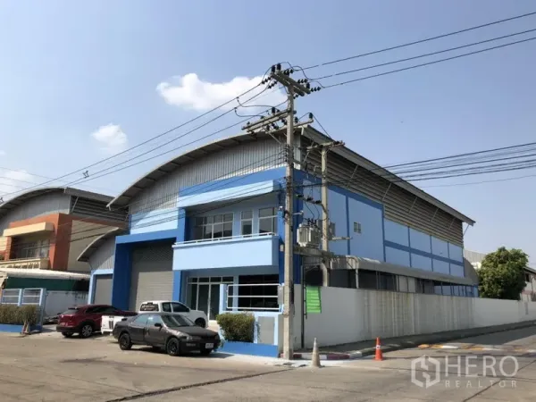 Factory for rent in Pathum Thani - Corner view of a blue warehouse building set on a wide industrial road.