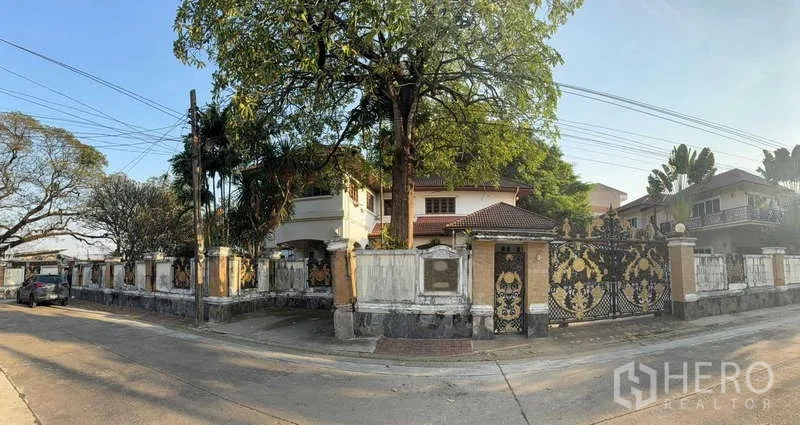 Detached House for sale in Sai Mai, Bangkok - Corner view of a large gated property with high perimeter wall, leafy shade trees and a decorative black-and-gold entrance.