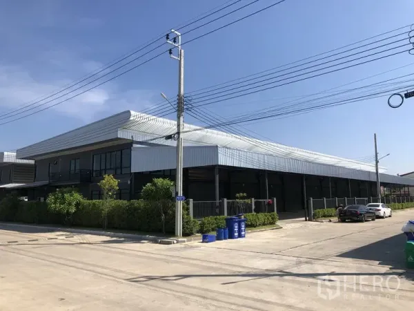 Factory for rent or sale in Bang Bo, Samut Prakan - Corner view of a modern steel-clad factory with wide concrete road and parking.