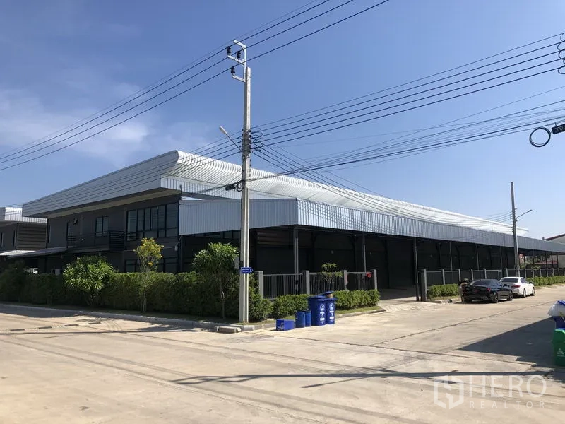 Factory for rent or sale in Bang Bo, Samut Prakan - Corner view of a modern steel-clad factory with wide concrete road and parking.