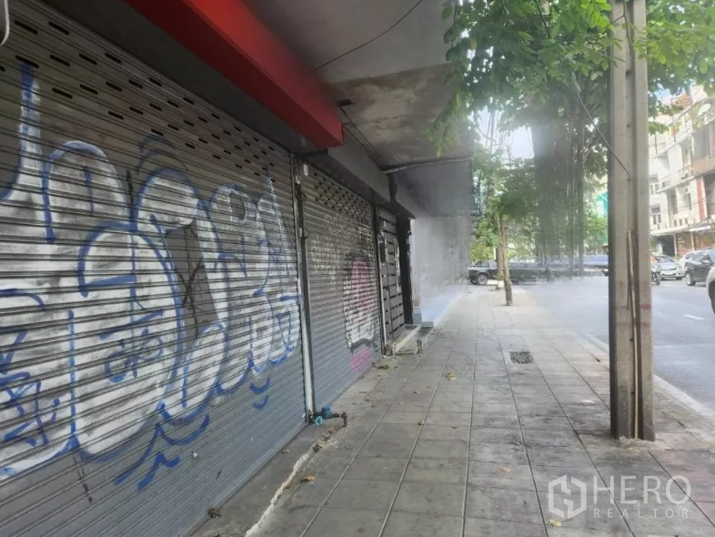 Shophouse for rent in Watthana, Bangkok - Sidewalk view of multiple shuttered shopfronts under shade trees on a main road.