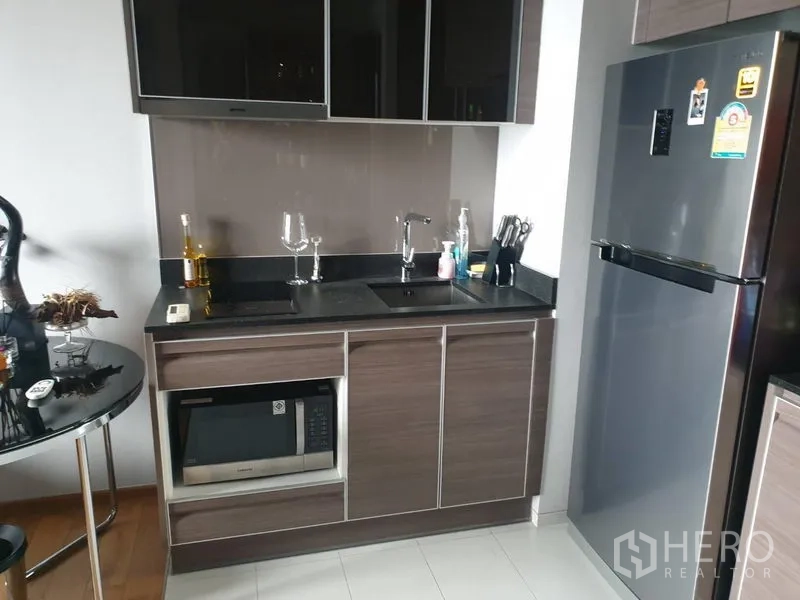Condominium for rent in Khlong Toei, Bangkok - Compact modern kitchenette with glossy cabinets, black countertop, sink, microwave, and stainless-steel refrigerator.