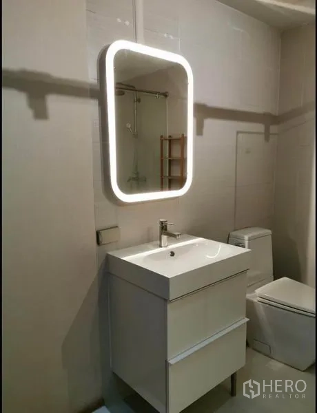 Condominium for rent in Khlong Toei, Bangkok - Vanity unit with backlit mirror and toilet in a bright tiled bathroom.
