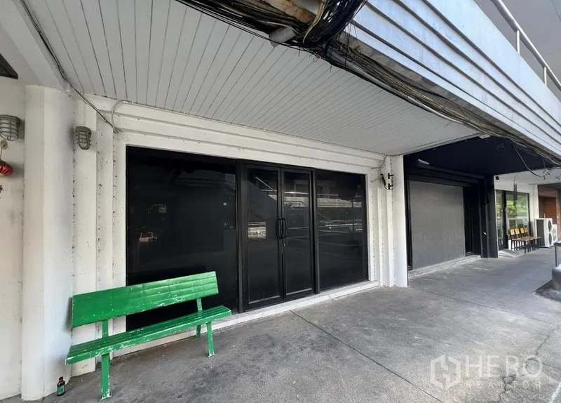 Shophouse for rent in Watthana, Bangkok - Exterior walkway with black glass storefront and a green bench along the facade.