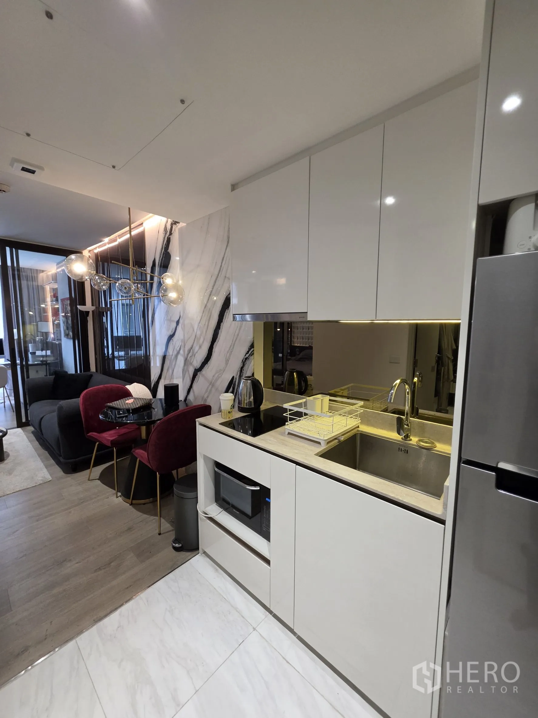 Condominium for rent in Khlong Toei, Bangkok - Compact white kitchen with sink, cooktop, microwave, and fridge next to living and dining areas.