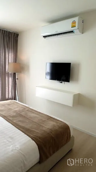 Condominium for sale in Khlong Toei, Bangkok - Bedroom wall-mounted TV beneath an air conditioner, with neutral decor and soft lighting.