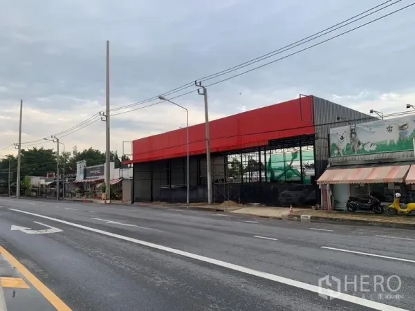 Warehouse for rent in Bueng Kum, Bangkok - Angled view of a roadside warehouse with red cladding, multi-lane road, streetlights and nearby shops.