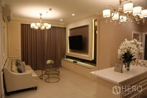 คอนโด ให้เช่าในHuai Khwang, Bangkok - ห้องนั่งเล่นโทนอบอุ่น พร้อมโซฟาสีเบจ โคมระย้า ผนังทีวีบิวท์อิน โต๊ะกลางกระจก ผ้าม่านทึบ และเคาน์เตอร์วางแจกันดอกไม้