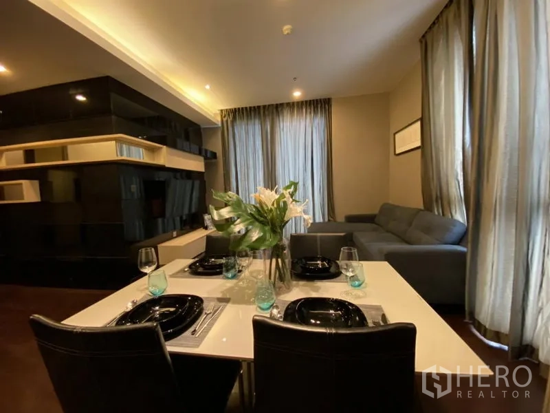 公寓 出租在Watthana, Bangkok - 开放式客餐厅，摆放四人餐桌、转角沙发、内置书架与落地窗帘。