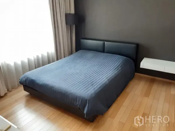 Condominium for rent in Khlong Toei, Bangkok - Spacious bedroom featuring a large bed, dark feature wall, wood floors and sheer curtains by the window.