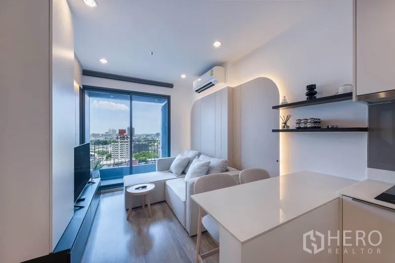 公寓 出租在Bang Na, Bangkok - 明亮客厅，L形沙发、电视柜，落地推拉门通向阳台俯瞰城市景观。