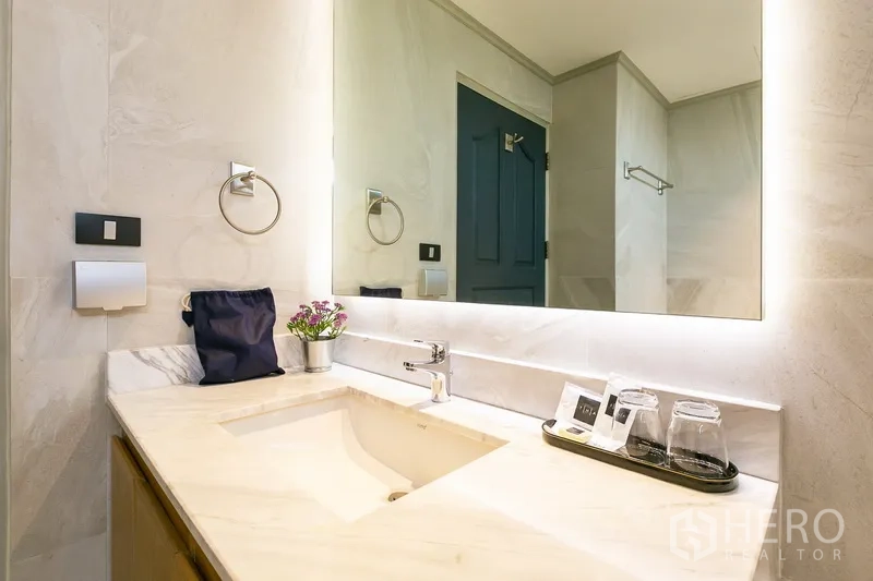 Apartment for rent in Khlong Toei, Bangkok - Bathroom vanity with backlit mirror, stone countertop and sink.