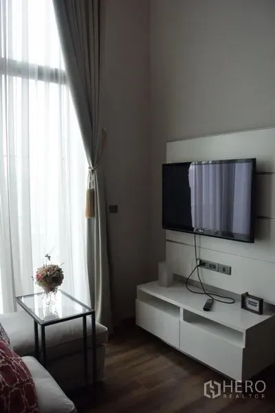 公寓 出租在Watthana, Bangkok - 墙挂电视与白色电视柜，旁有高窗与纱帘。