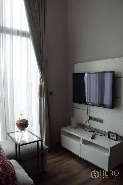 公寓 出租在Watthana, Bangkok - 墙挂电视与白色电视柜，旁有高窗与纱帘。