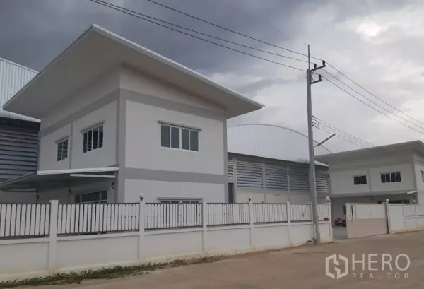 Factory for rent in Nakhon Pathom - Modern standalone factory with two-story office, white perimeter fence and wide road in an industrial compound under cloudy skies.