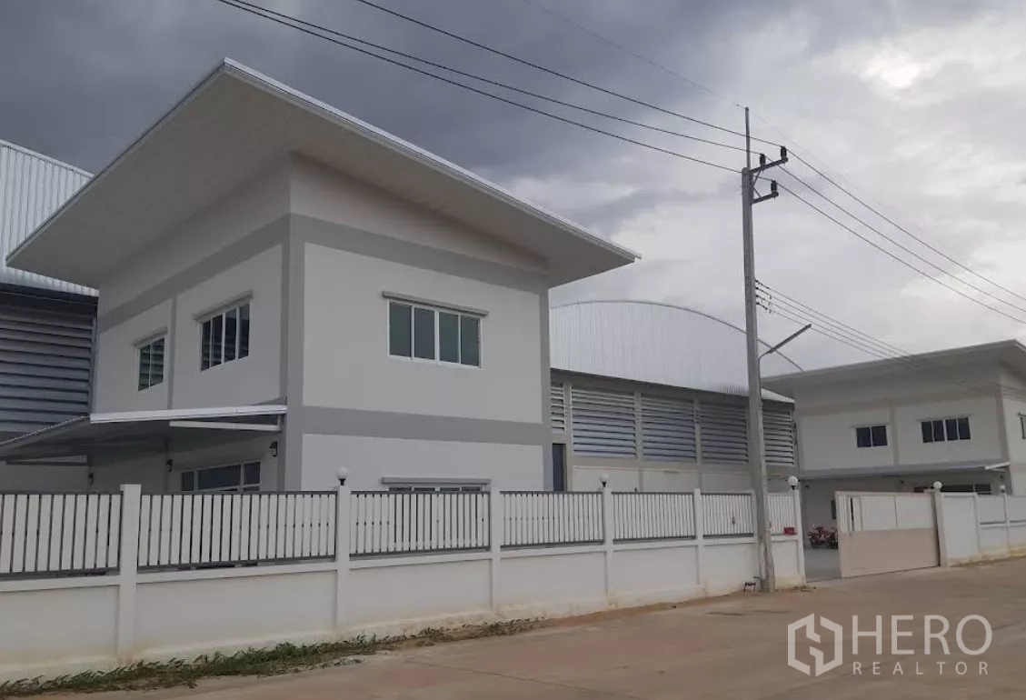 Factory for rent in Nakhon Pathom - Modern standalone factory with two-story office, white perimeter fence and wide road in an industrial compound under cloudy skies.