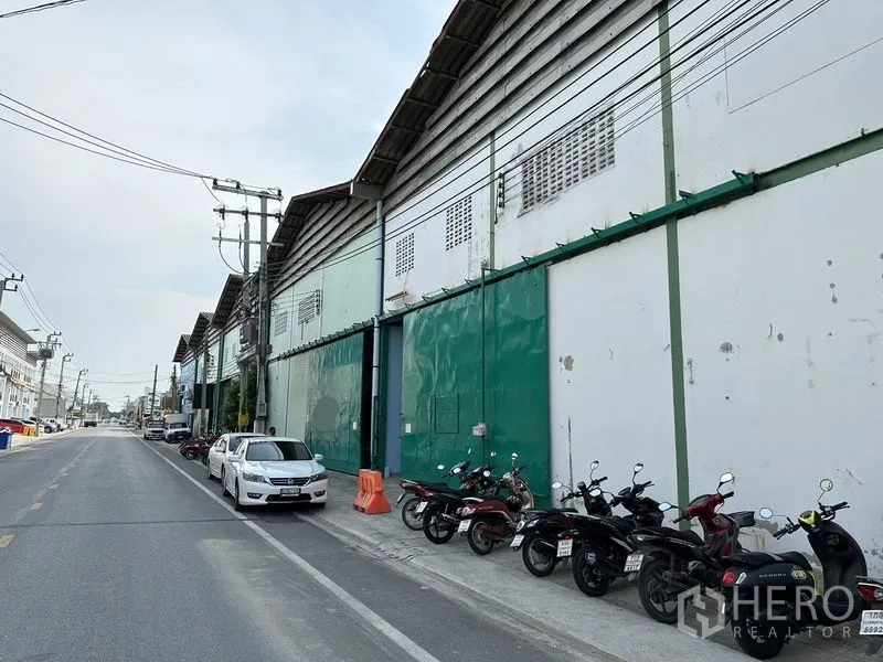 Factory for rent in Bang Bo, Samut Prakan - Wider streetscape showing a row of warehouse-style factories with green doors and wide road access.