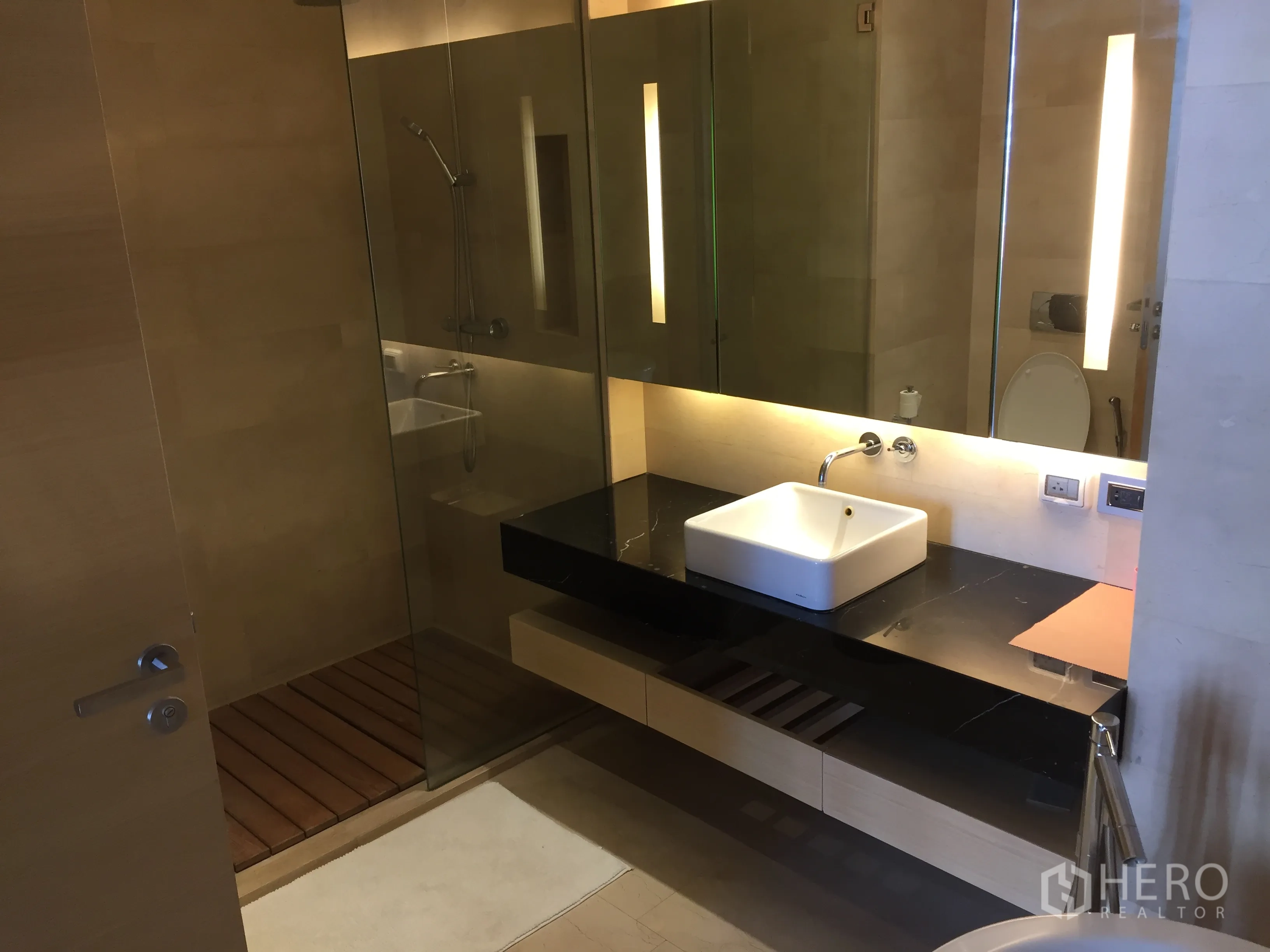 Condominium for rent or sale in Bang Rak, Bangkok - Glass-enclosed walk-in shower beside a long vanity with illuminated mirror.