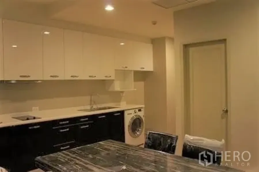 Condominium for rent in Watthana, Bangkok - Modern condo kitchen with white upper cabinets, black base cabinets, sink, cooktop and a front‑load washer beside a marble dining table.