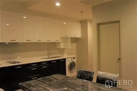 Condominium for rent in Watthana, Bangkok - Modern condo kitchen with white upper cabinets, black base cabinets, sink, cooktop and a front‑load washer beside a marble dining table.