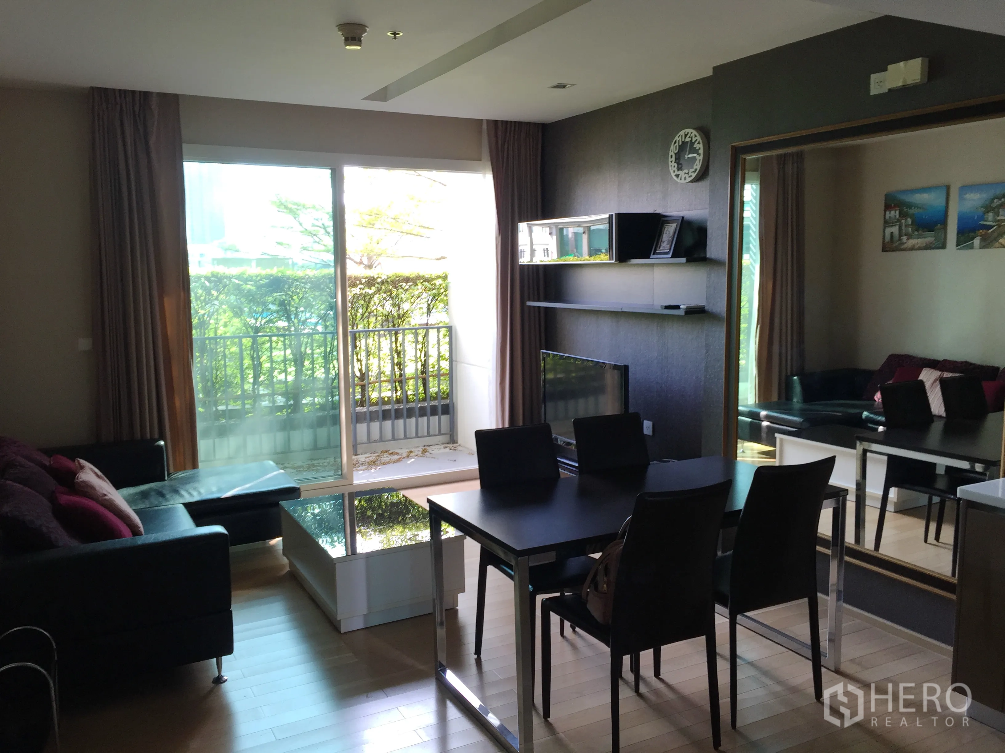 Condominium for rent in Khlong Toei, Bangkok - Open-plan living and dining area with sofa, dining table, TV wall and balcony facing greenery.
