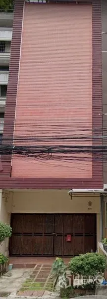 Shophouse for rent in Khlong Toei, Bangkok - Four-story shophouse exterior with full-height pink roller shutter, wooden entry doors, overhead utility cables, and a small front garden.