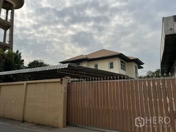 Detached House for rent or sale in Lak Si, Bangkok - Street view of tall sliding gate and beige wall with carport roof and a two-storey house under a cloudy sky.