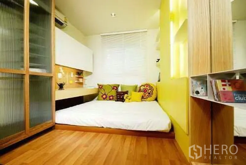 Condominium for rent in Khlong San, Bangkok - Cozy bedroom with platform bed, yellow accent wall, shelving and a frosted-glass wardrobe.