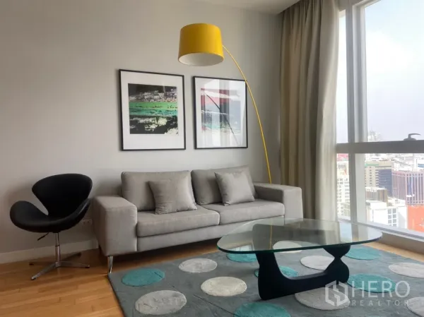 Condominium for rent or sale in Khlong Toei, Bangkok - Cozy seating area with grey sofa, modern art, patterned rug and large window overlooking Bangkok.