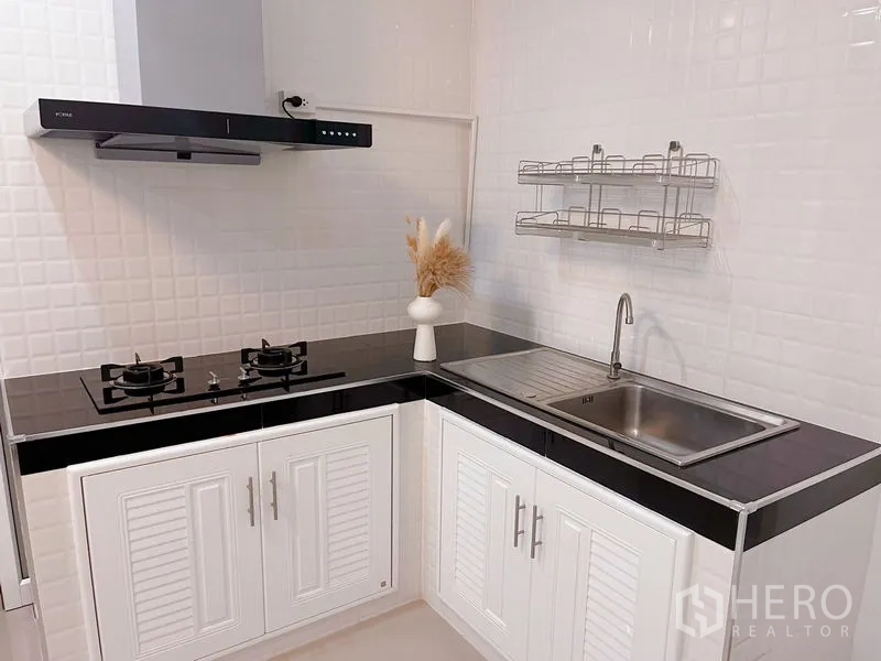 Townhouse for rent in Huai Khwang, Bangkok - Modern L-shaped kitchen with gas hobs, hood, sink and white storage cabinets.