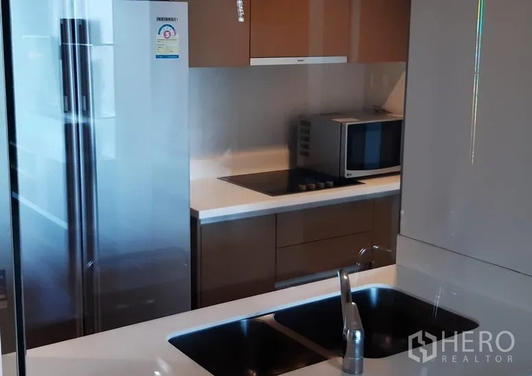 Condominium for rent in Khlong Toei, Bangkok - Modern condo kitchen with double sink, stainless refrigerator, microwave and electric cooktop with glossy cabinetry.