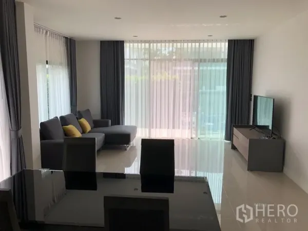 Detached House for rent in Bang Kapi, Bangkok - Sunlit living room with L-shaped sofa, floor-to-ceiling sheer curtains, and a TV console.