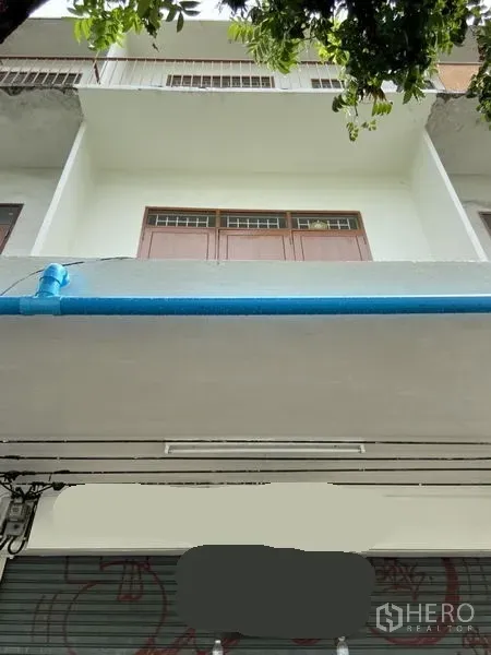 Shophouse for rent in Khlong Toei, Bangkok - Street-facing shophouse facade with upper balcony and roll-up shutter under tree shade.