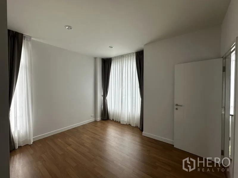 Detached House for sale in Lam Luk Ka, Pathum Thani - Sunlit bedroom with wood flooring and tall sheer-curtained windows.