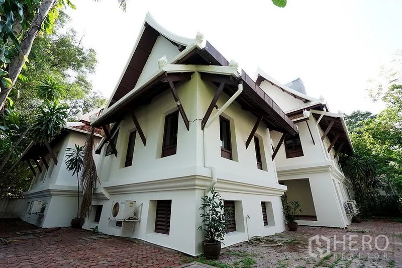 独立式住宅 出租在Watthana, Bangkok - 房屋侧立面展示特色飞檐、铺装庭院、空调外机与热带绿植。