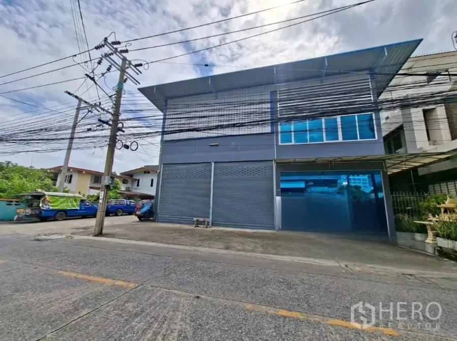 Warehouse for rent in Suan Luang, Bangkok - Street view of a two‑storey warehouse with gray facade, blue roof, roller shutters and a front driveway in Suan Luang.