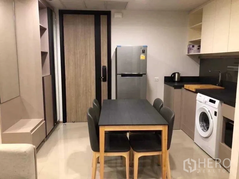 Condominium for rent in Khlong Toei, Bangkok - Open kitchen and dining area with four-seat table, fridge, washing machine, and built-in cabinetry.