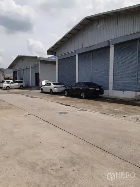 货栈 出租在Bang Phli, Samut Prakan - 一排带卷闸门的仓库建筑沿宽混凝土道路分布。