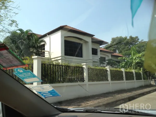 บ้าน ให้เช่าในNonthaburi - บ้านเดี่ยวสองชั้นสีขาว หลังคากระเบื้องสีน้ำตาล หน้าต่างบานใหญ่ และรั้วเหล็ก ล้อมด้วยต้นไม้ร่มรื่น ในปากเกร็ด นนทบุรี