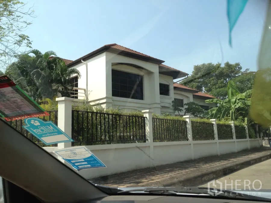 บ้าน ให้เช่าในNonthaburi - บ้านเดี่ยวสองชั้นสีขาว หลังคากระเบื้องสีน้ำตาล หน้าต่างบานใหญ่ และรั้วเหล็ก ล้อมด้วยต้นไม้ร่มรื่น ในปากเกร็ด นนทบุรี