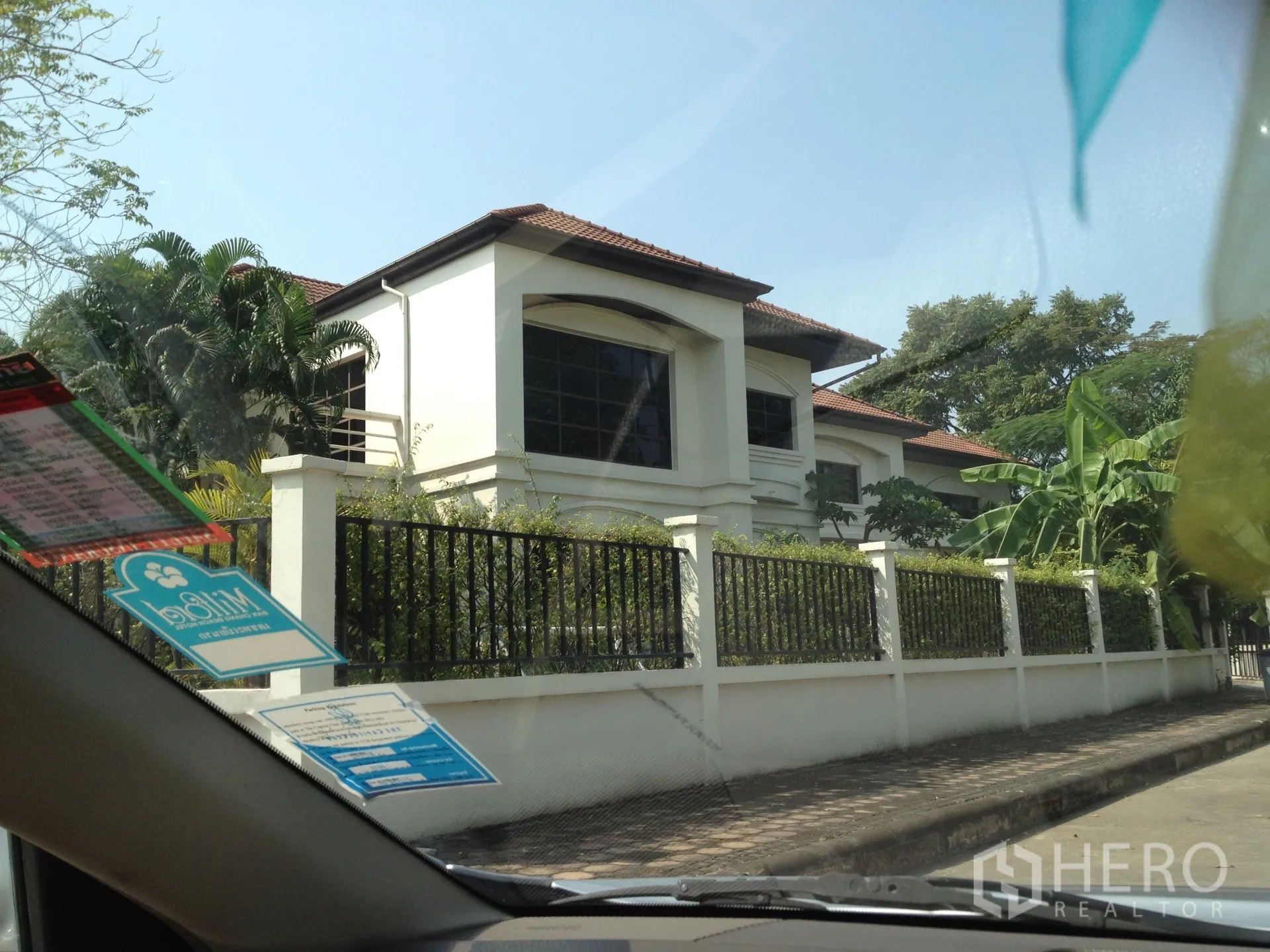 บ้าน ให้เช่าในNonthaburi - บ้านเดี่ยวสองชั้นสีขาว หลังคากระเบื้องสีน้ำตาล หน้าต่างบานใหญ่ และรั้วเหล็ก ล้อมด้วยต้นไม้ร่มรื่น ในปากเกร็ด นนทบุรี