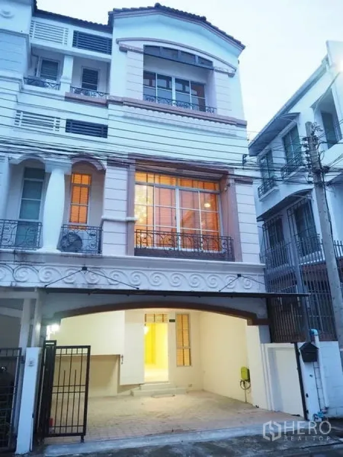Townhouse for rent in Lat Phrao, Bangkok - Three-storey townhouse exterior at dusk with large windows, balconies and a gated carport in Lat Phrao, Bangkok.